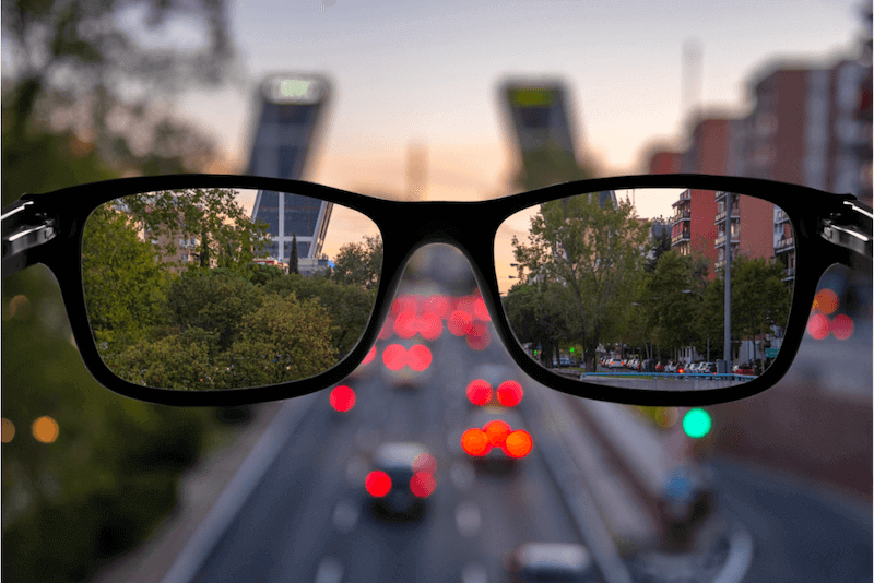 Óculos de grau visto de perto com uma cidade e trânsito ao fundo, refletindo um cenário urbano ao entardecer.