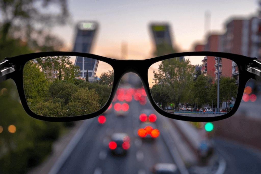 Óculos de grau visto de perto com uma cidade e trânsito ao fundo, refletindo um cenário urbano ao entardecer