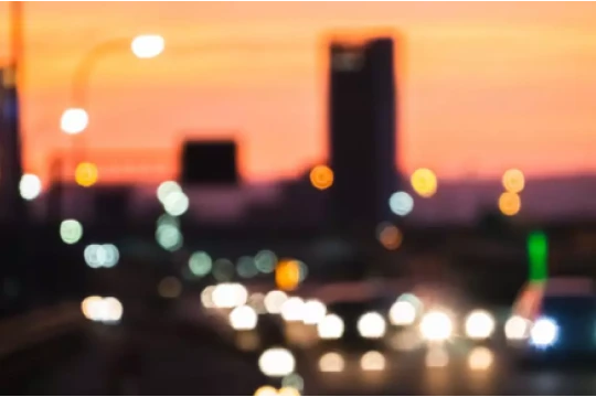 Imagem de uma cidade ao entardecer, com luzes de carros em movimento em uma estrada. O céu apresenta tonalidades de laranja e roxo, criando uma atmosfera urbana vibrante.