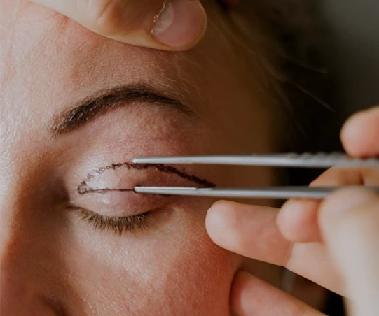 Aplicação de maquiagem com delineador em um olho feminino, onde um profissional usa pinças para criar um traço detalhado.