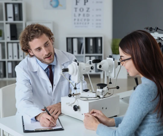 Um oftalmologista conversando com uma paciente em um consultório, com equipamentos oftalmológicos no ambiente.