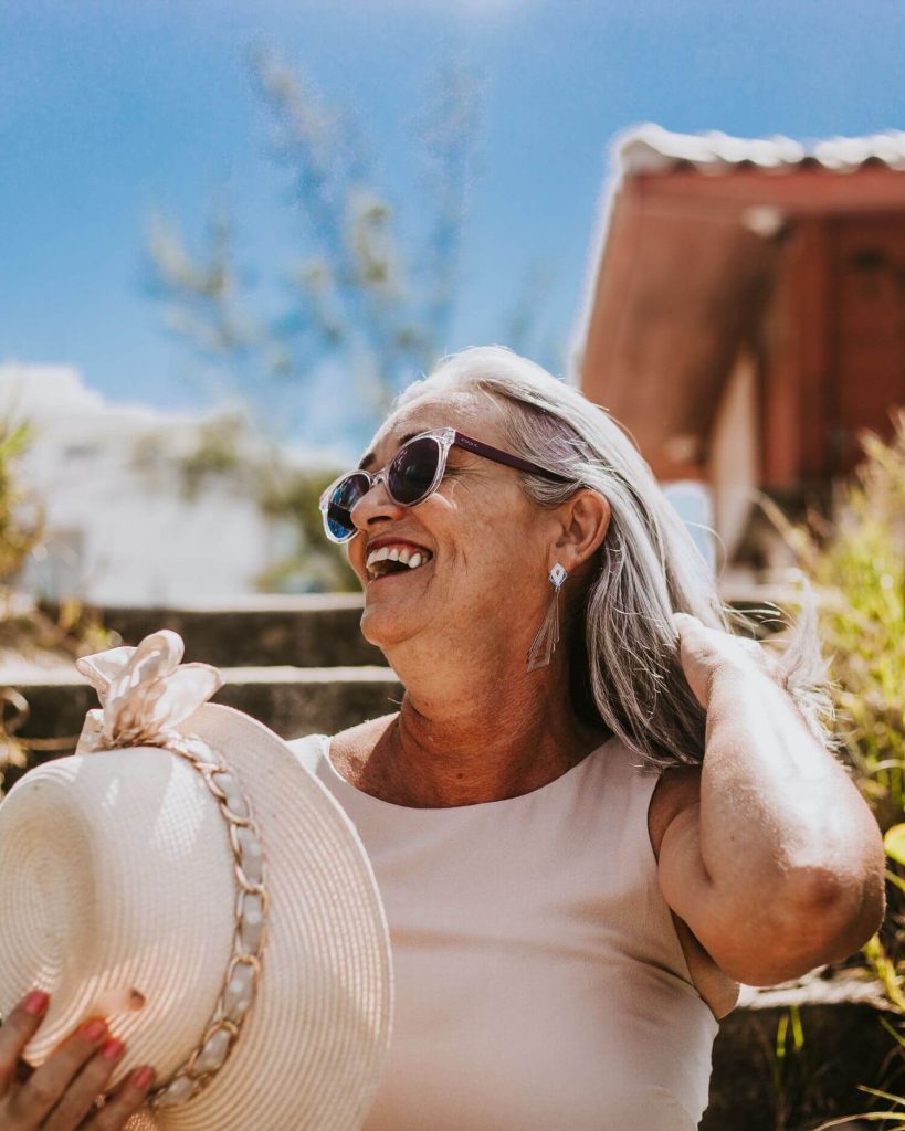 Mulher idosa sorrindo ao ar livre, usando óculos de sol e chapéu de palha, desfrutando de um dia ensolarado em um ambiente natural com escadas de madeira e vegetação ao fundo.